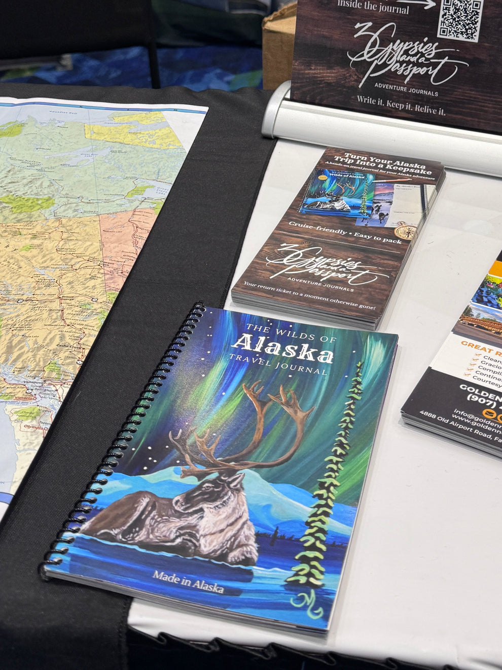 A tabletop display of printed Alaska-themed brochures, a spiral notebook, and a standing sign at what appears to be a booth or exhibition. The materials feature scenic Alaska photography, maps, and promotional text, with chairs and other booth elements visible in the background.