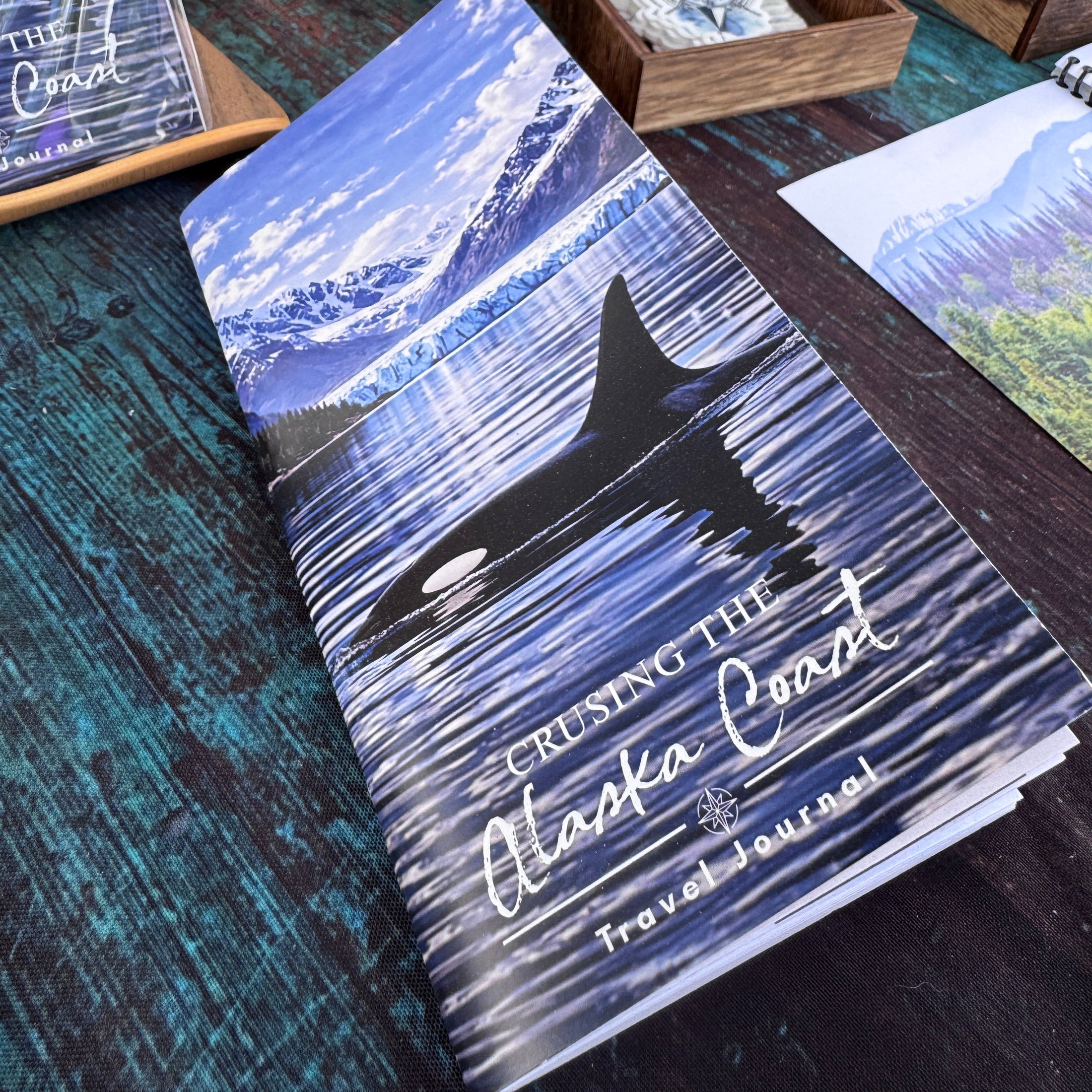 Crusing the Alaska Coast Travel Journal with an orca swimming on it on a table with other items in the background