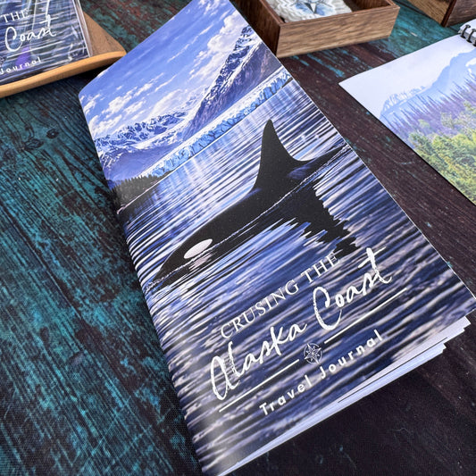 Crusing the Alaska Coast Travel Journal with an orca swimming on it on a table with other items in the background