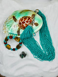 Load image into Gallery viewer, Turquoise colored beaded necklace on turtle jewelry box in the sand with a bracelet and ring
