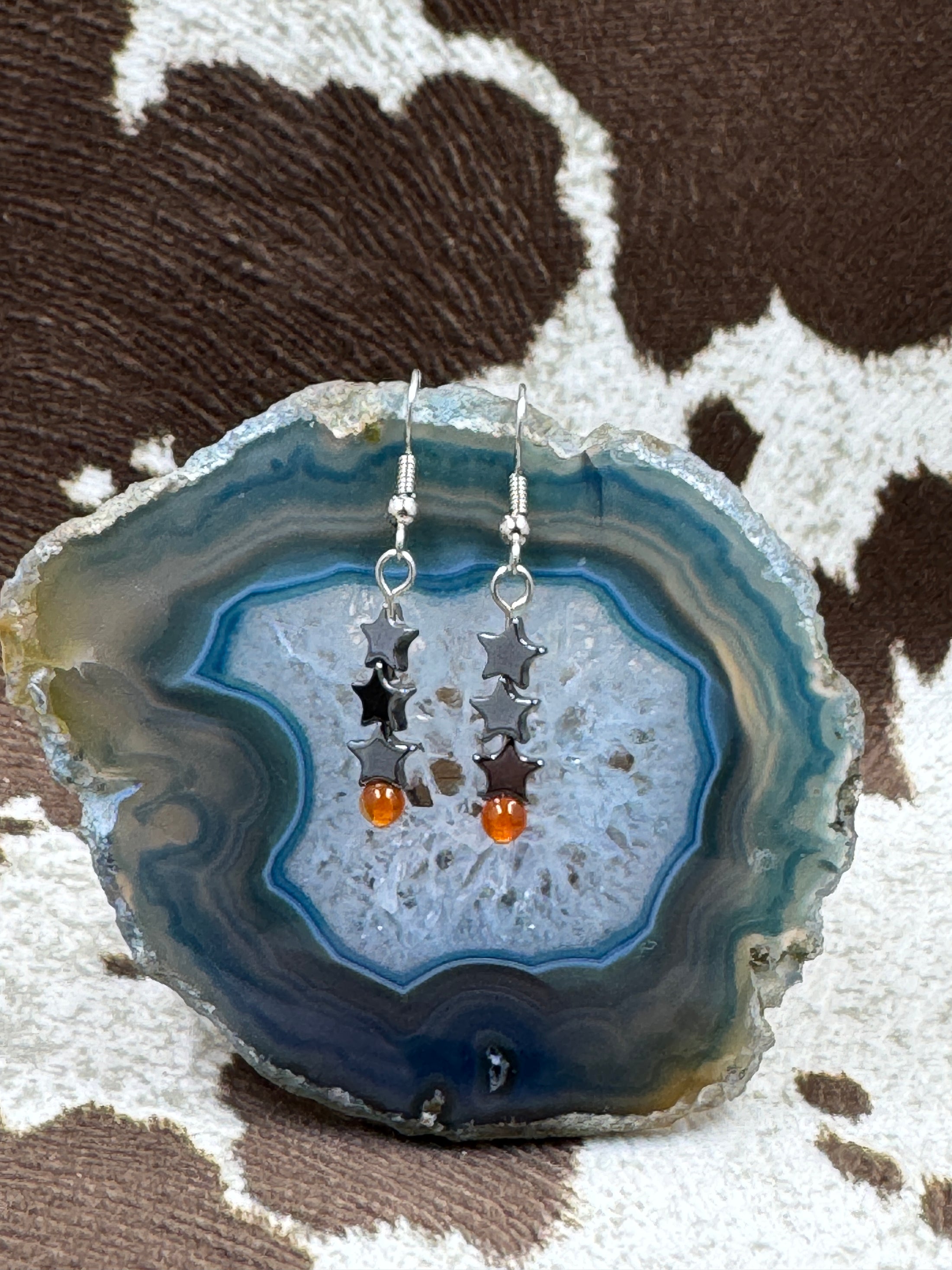 Hematite and Carnelian Star Earrings hanging from a blue stone rock with cowhide background