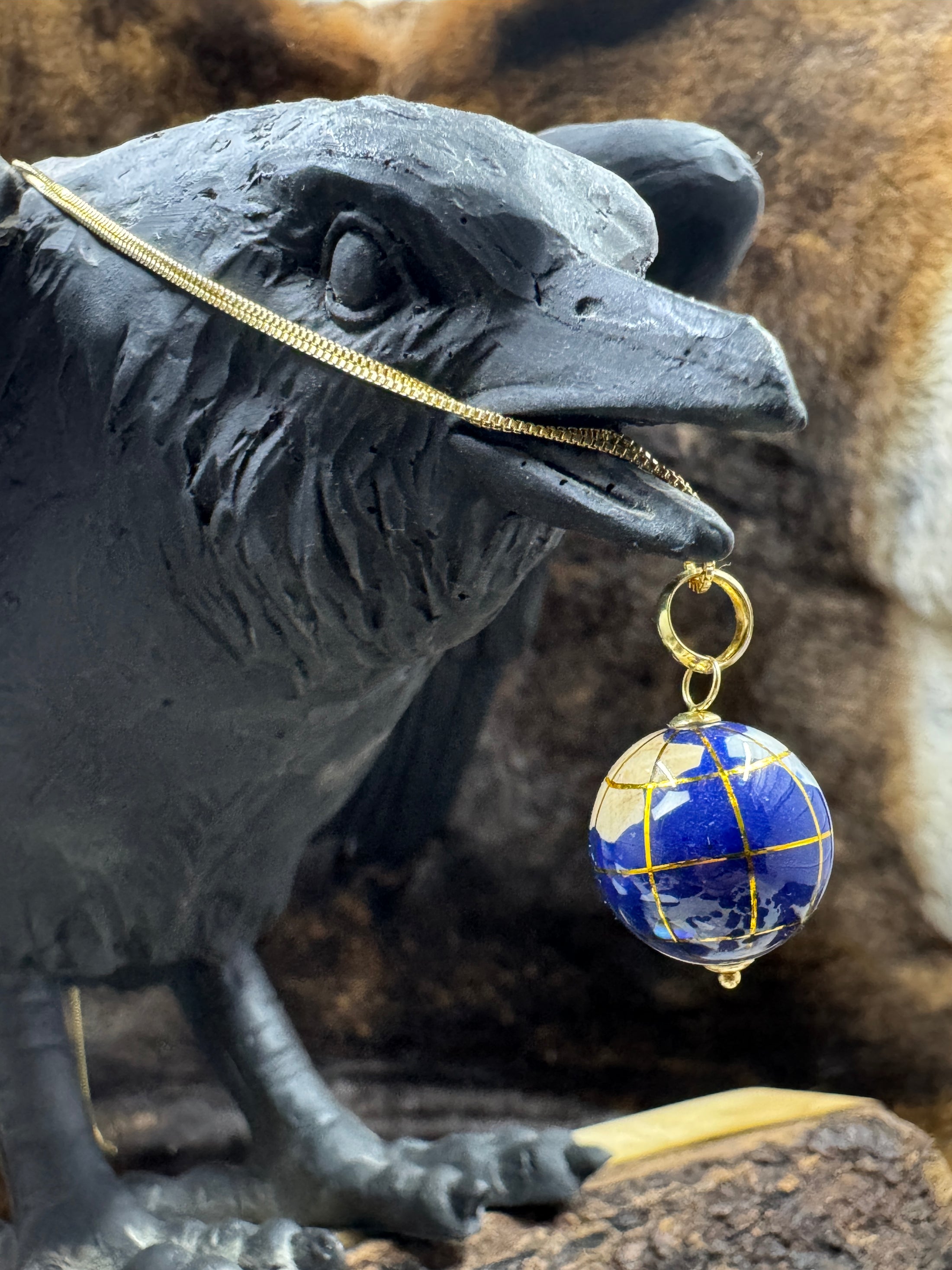 Vintage 14K World Glob Inlay Charm Pendant by Michael Anthony hanging on a gold chain from raven statue with fur background