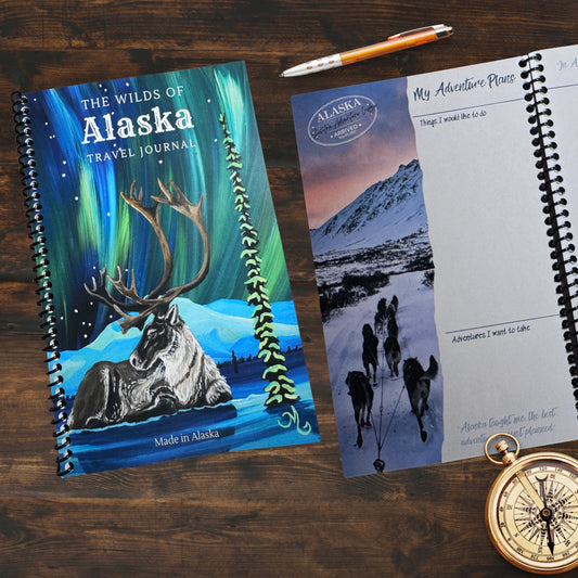 The wilds of Alaska Travel Journal cover and open journal laying on desk with a pen and compass