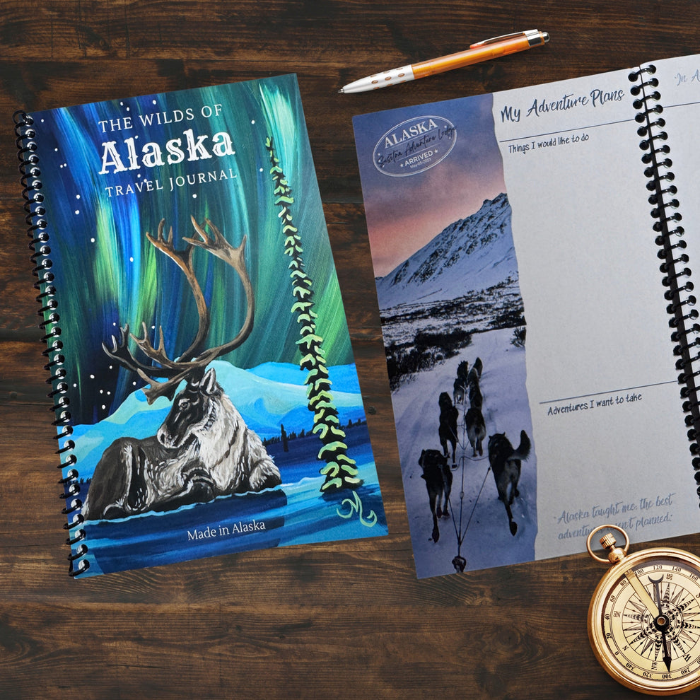 The wilds of Alaska Travel Journal cover and open journal laying on desk with a pen and compass